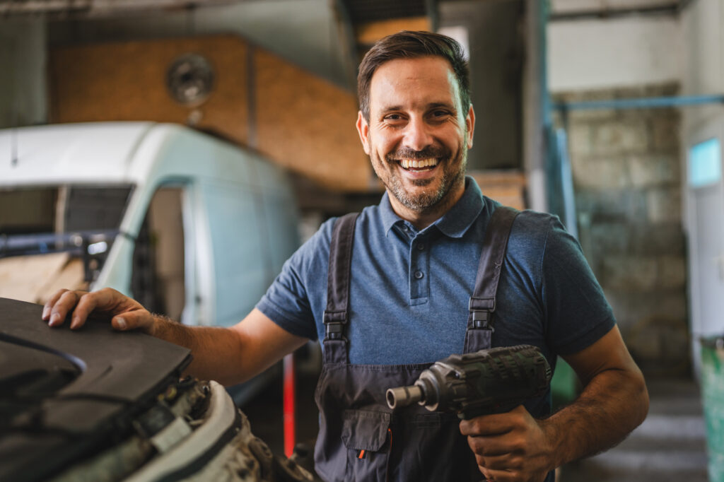 smiling mechanic holding a drill and standing beside a vehicle inside a shop